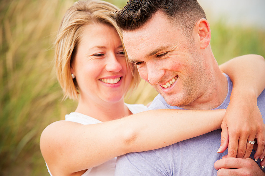 Couple smiling during their portrait session on the beach, with tall grass on the dunes behind them.
