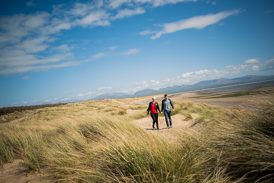 Photograph of Liz & Jason walking through the sand dunes, holding hands. Photographed by Celynnen Photography.